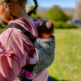 Back of child carrying a doll in a Didymos toy carrier in olive twig