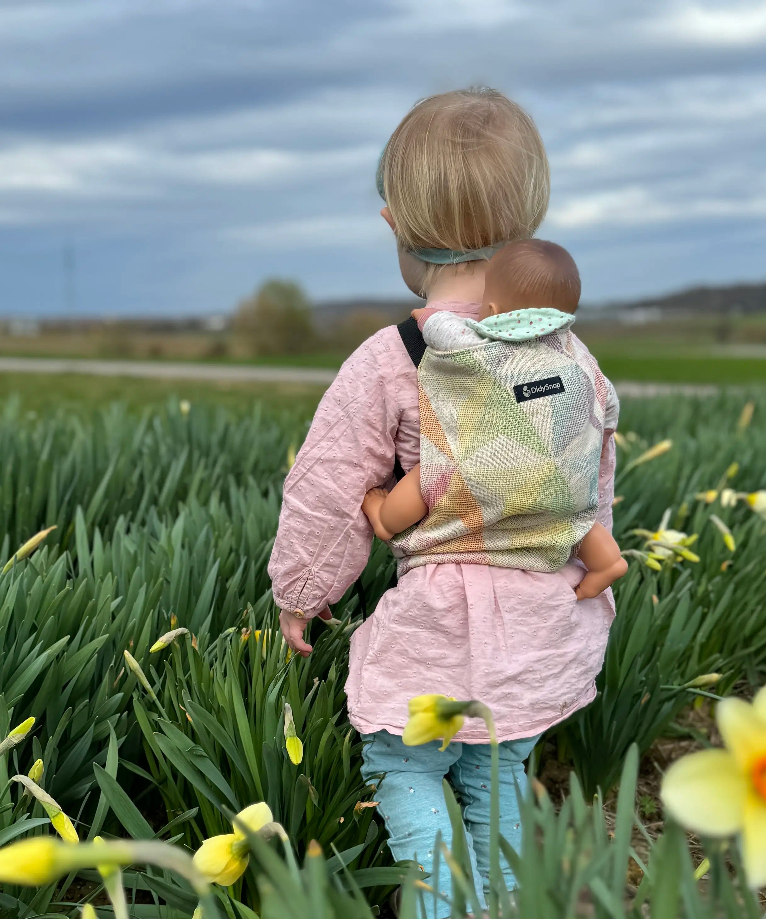 Child carrying a doll in a Didymos toy carrier in the zephyr design in a grassy field