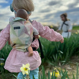 Back of child carrying a doll in a Didymos toy carrier in the zephyr design. A cream carrier with repeating geometric shapes in a pastel palette