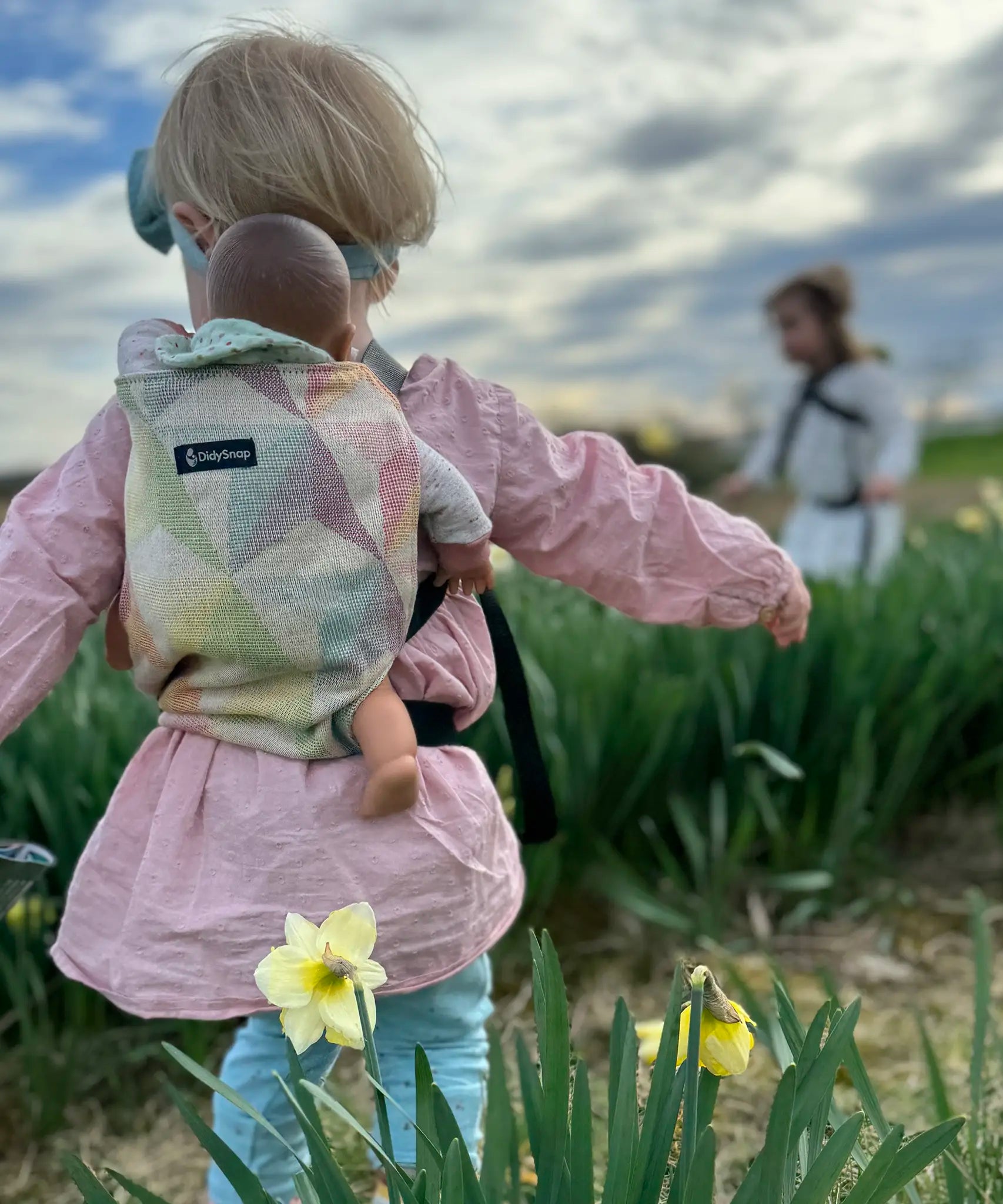 Back of child carrying a doll in a Didymos toy carrier in the zephyr design. A cream carrier with repeating geometric shapes in a pastel palette
