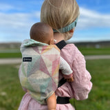Child carrying a doll in a Didymos toy cotton carrier in the zephyr. A cream carrier with repeating geometric shapes in a pastel palette