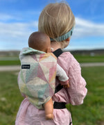 Child carrying a doll in a Didymos toy cotton carrier in the zephyr. A cream carrier with repeating geometric shapes in a pastel palette