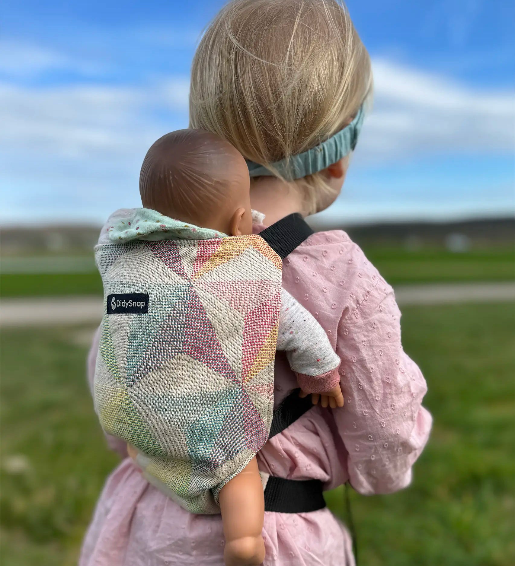 Child carrying a doll in a Didymos toy cotton carrier in the zephyr. A cream carrier with repeating geometric shapes in a pastel palette