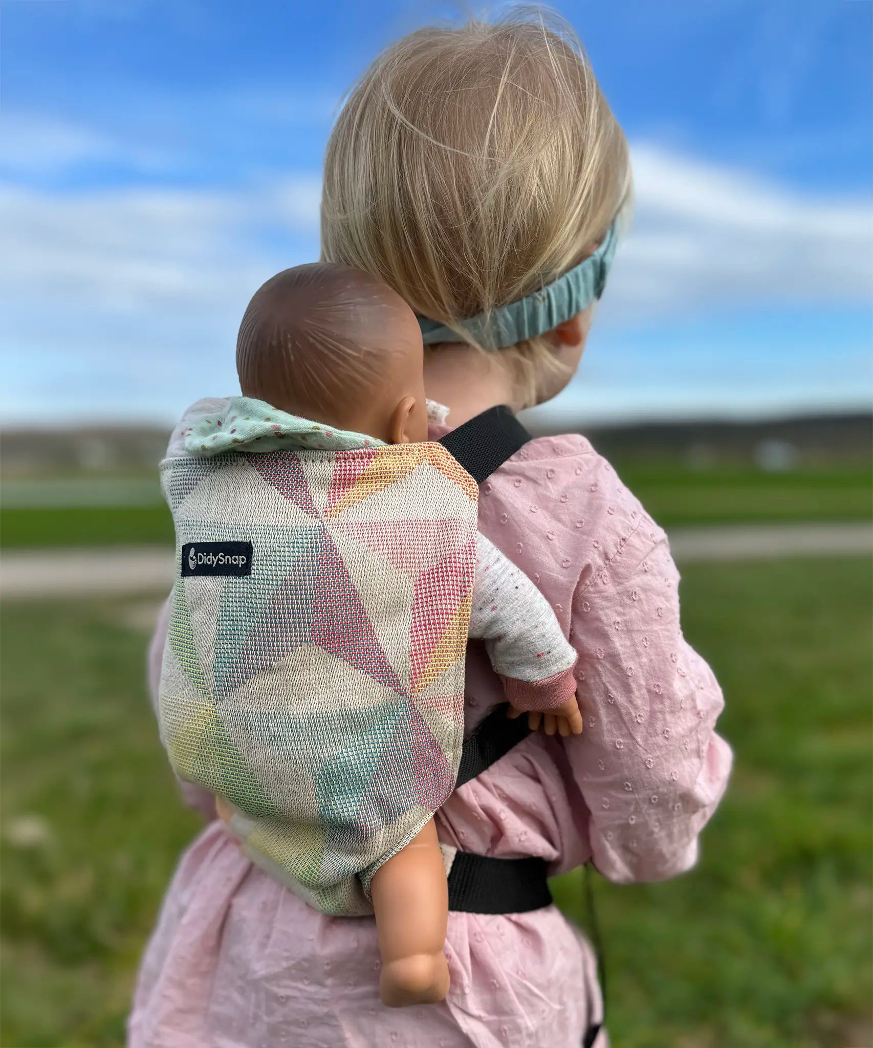 Child carrying a doll in a Didymos toy cotton carrier in the zephyr. A cream carrier with repeating geometric shapes in a pastel palette
