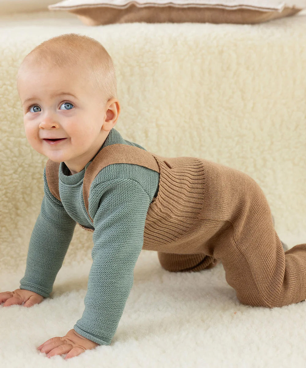 Baby crawling indoors wearing caramel brown Disana knitted merino wool dungarees over a long-sleeved top.
