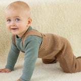 Baby crawling indoors wearing caramel brown Disana knitted merino wool dungarees over a long-sleeved top.