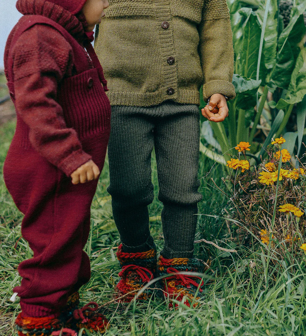 Toddler wearing deep maroon knitted merino wool dungarees and matching wool layers by Disana, standing in grass outdoors.