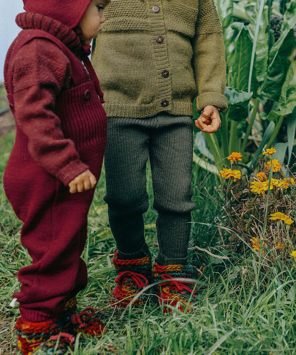 Toddler wearing deep maroon knitted merino wool dungarees and matching wool layers by Disana, standing in grass outdoors.