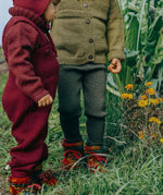 Toddler wearing deep maroon knitted merino wool dungarees and matching wool layers by Disana, standing in grass outdoors.