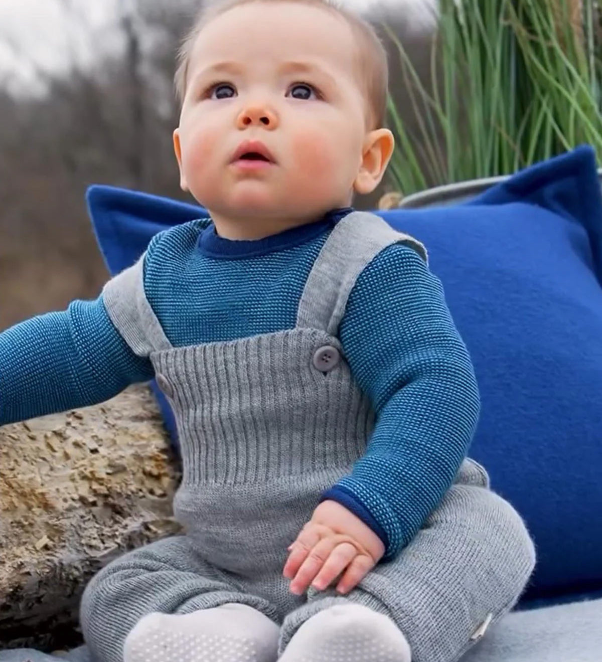 Baby wearing light grey Disana knitted merino wool dungarees with buttoned straps, seated outdoors on a blanket.
