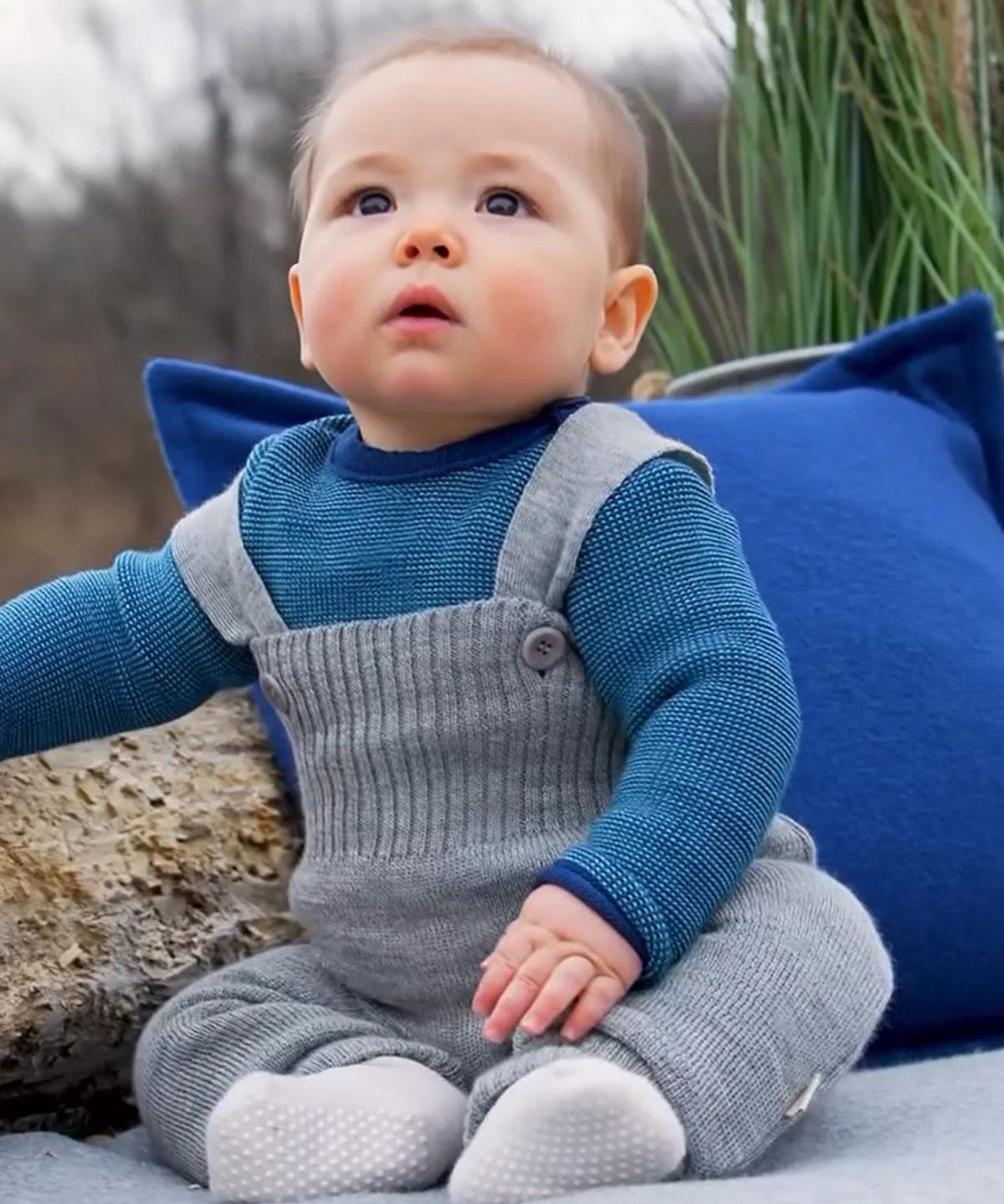 Baby wearing light grey Disana knitted merino wool dungarees with buttoned straps, seated outdoors on a blanket.