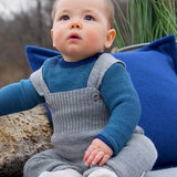 Baby wearing light grey Disana knitted merino wool dungarees with buttoned straps, seated outdoors on a blanket.