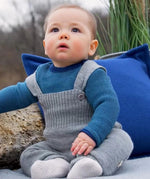 Baby wearing light grey Disana knitted merino wool dungarees with buttoned straps, seated outdoors on a blanket.