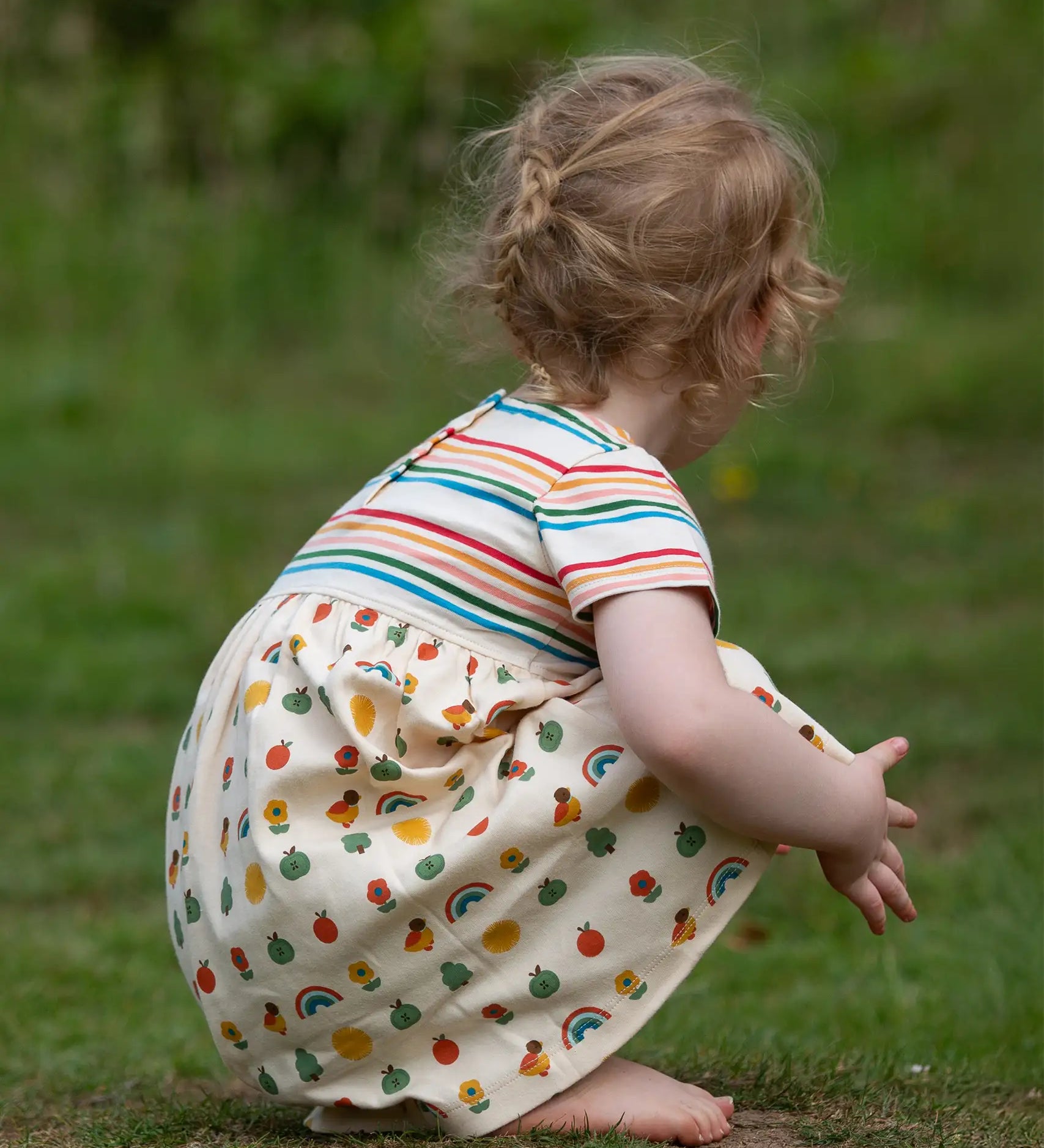 Crouching child wearing the Little Green Radicals Easy Peasy organic cotton dress with a cream rainbow striped body and nature patterned skirt.
