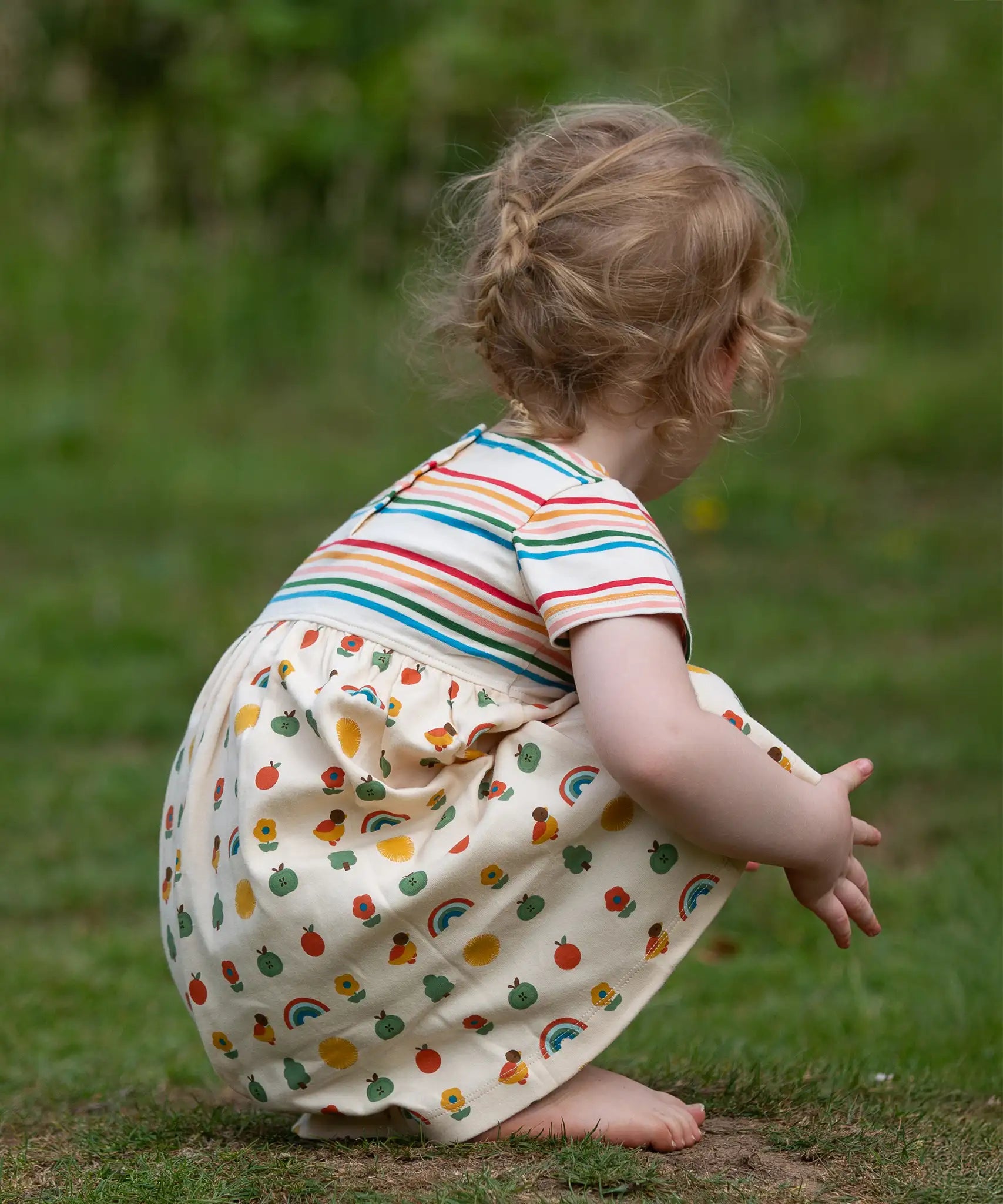 Crouching child wearing the Little Green Radicals Easy Peasy organic cotton dress with a cream rainbow striped body and nature patterned skirt.