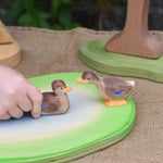 Two Ostheimer wooden ducks placed on a wooden pond scene piece. A child's hand can be seen holding the swimming duck.