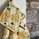DUNS Sweden organic cotton linen pot holder and tea towel in the bee yellow print hanging from a metal hook in front of a stone wall