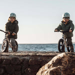 2 children sat on Early Rider aluminium balance bikes on a rock in front of the sea