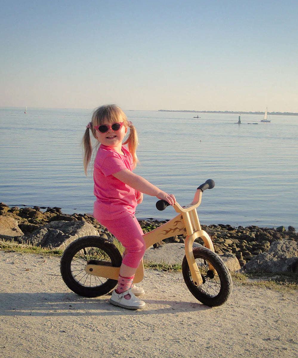 A little girl riding The Early Rider Lite Wooden Balance Bike with the ocean in the background