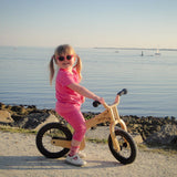 A little girl riding The Early Rider Lite Wooden Balance Bike with the ocean in the background