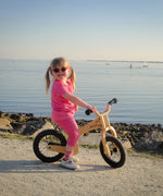 A little girl riding The Early Rider Lite Wooden Balance Bike with the ocean in the background