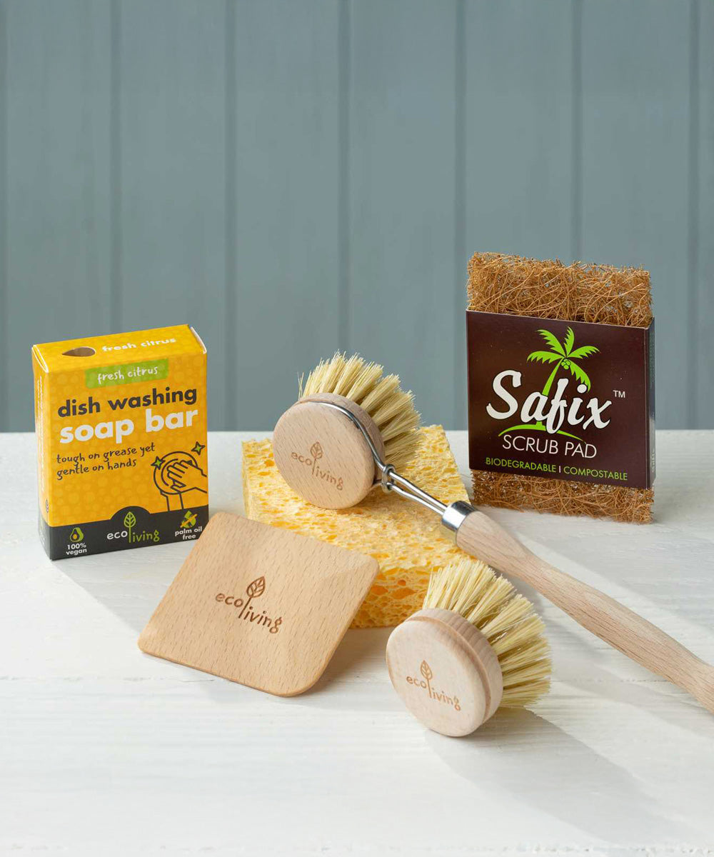 The content of the EcoLiving washing up starter set placed on a white work surface. The set consists of a coconut soap rest, a reusable sponge a wooden dish brush & replacement head, dish soap and a wooden pot scraper. 