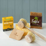 The content of the EcoLiving washing up starter set placed on a white work surface. The set consists of a coconut soap rest, a reusable sponge a wooden dish brush & replacement head, dish soap and a wooden pot scraper. 