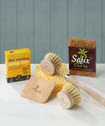 The content of the EcoLiving washing up starter set placed on a white work surface. The set consists of a coconut soap rest, a reusable sponge a wooden dish brush & replacement head, dish soap and a wooden pot scraper. 