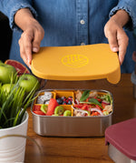 An adults hands lifting the lid off a Eco Rascals Stainless Steel Lunch Box with a mustard yellow silicone lid and pot inserts. The lunchbox is filled with food.