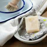 Lathered blossom breeze Ecoliving 100% vegan dish soap bar in a small bowl with a kitchen items in the background.