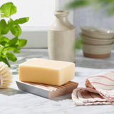 Ecoliving beech wood soap dish with a cream soap on top on a grey marble work top next to a tea towel and a green plant