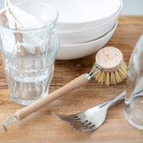 A short handled wooden dish brush placed on a wooden work surface surrounded by piles of white dishes and glassware. 