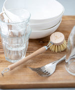 A short handled wooden dish brush placed on a wooden work surface surrounded by piles of white dishes and glassware. 