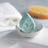 Close up of an Ecoliving solid sample size shampoo bar in a white soap dish on a sink