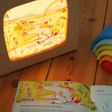 The river artwork by Stephanie Green  from the Toverlux Edulux water cycles set shown in a ToverLux Magic lamp. The accompanying booklet is open on the pages about the river in front of the lamp. A range of Toverlux silhouettes and lamps are available at Babipur.