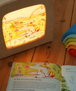 The river artwork by Stephanie Green  from the Toverlux Edulux water cycles set shown in a ToverLux Magic lamp. The accompanying booklet is open on the pages about the river in front of the lamp. A range of Toverlux silhouettes and lamps are available at Babipur.