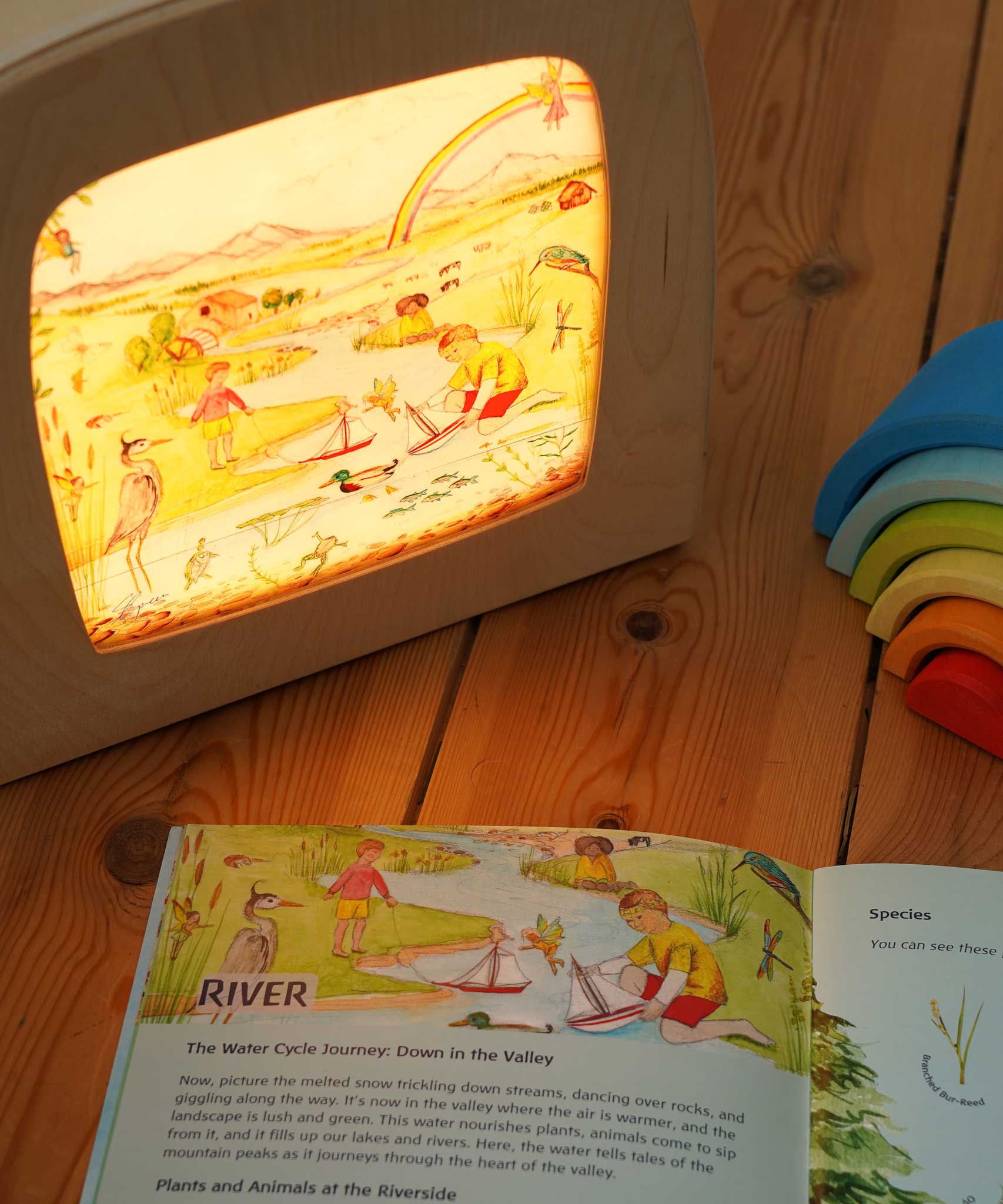 The river artwork by Stephanie Green  from the Toverlux Edulux water cycles set shown in a ToverLux Magic lamp. The accompanying booklet is open on the pages about the river in front of the lamp. A range of Toverlux silhouettes and lamps are available at Babipur.