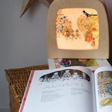 Around The Year children's hardback book by Elsa Beskow shown open on the August page with a Toverlux lamp with August artwork displayed in the background .