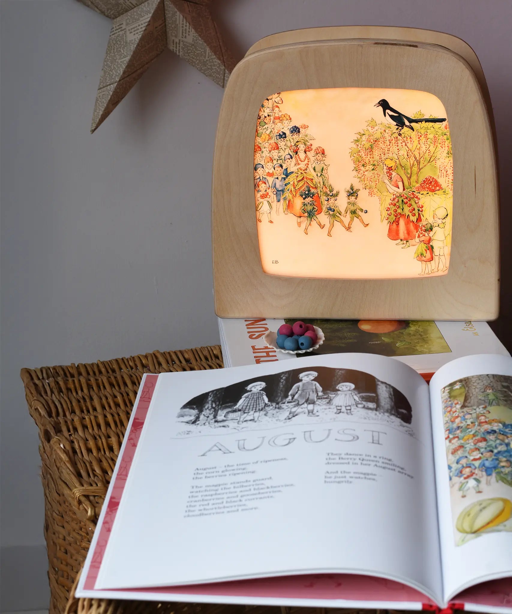 Around The Year children's hardback book by Elsa Beskow shown open on the August page with a Toverlux lamp with August artwork displayed in the background .