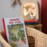 Around The Year children's hardback book by Elsa Beskow placed on a child's wicker chair with a Toverlux lamp with December artwork displayed in the background with a decorated celebration ring.