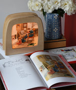 Around The Year children's hardback book by Elsa Beskow shown open on the November page with a Toverlux lamp with November artwork displayed in the background.