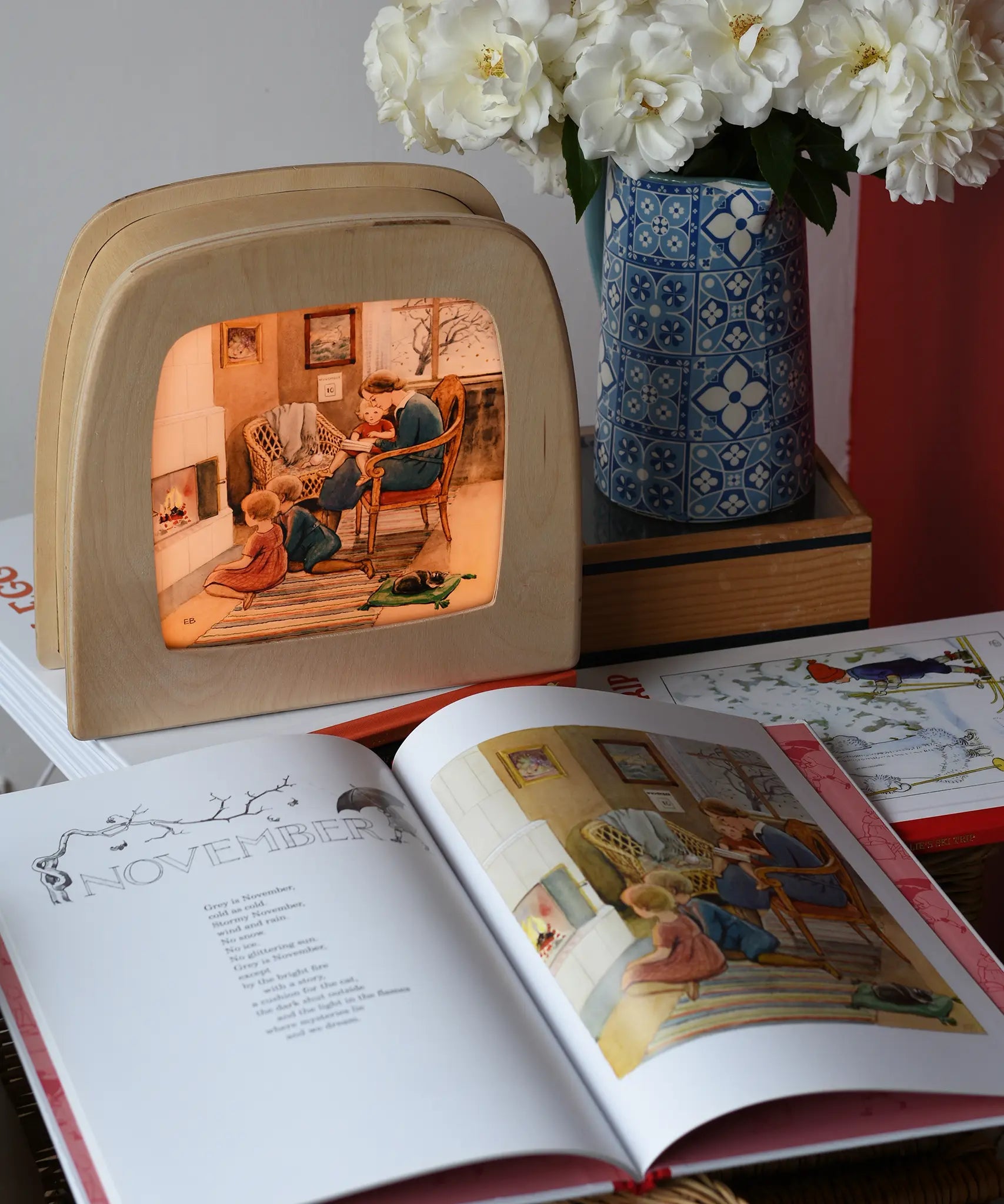 Around The Year children's hardback book by Elsa Beskow shown open on the November page with a Toverlux lamp with November artwork displayed in the background.