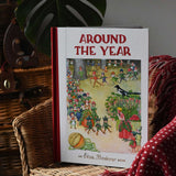 Around The Year children's hardback book by Elsa Beskow placed on a child's rattan chair next to a red blanket.