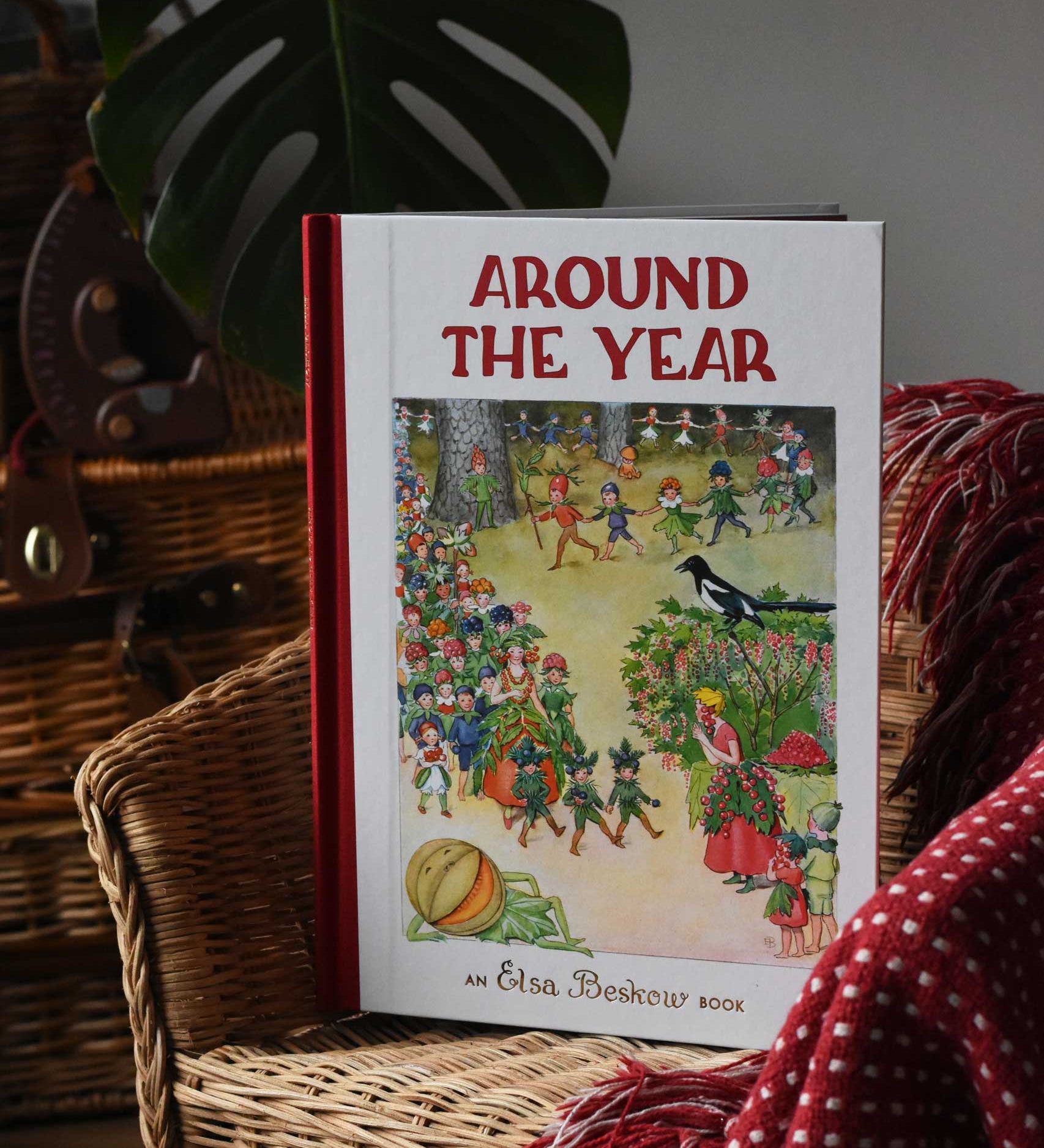 Around The Year children's hardback book by Elsa Beskow placed on a child's rattan chair next to a red blanket.