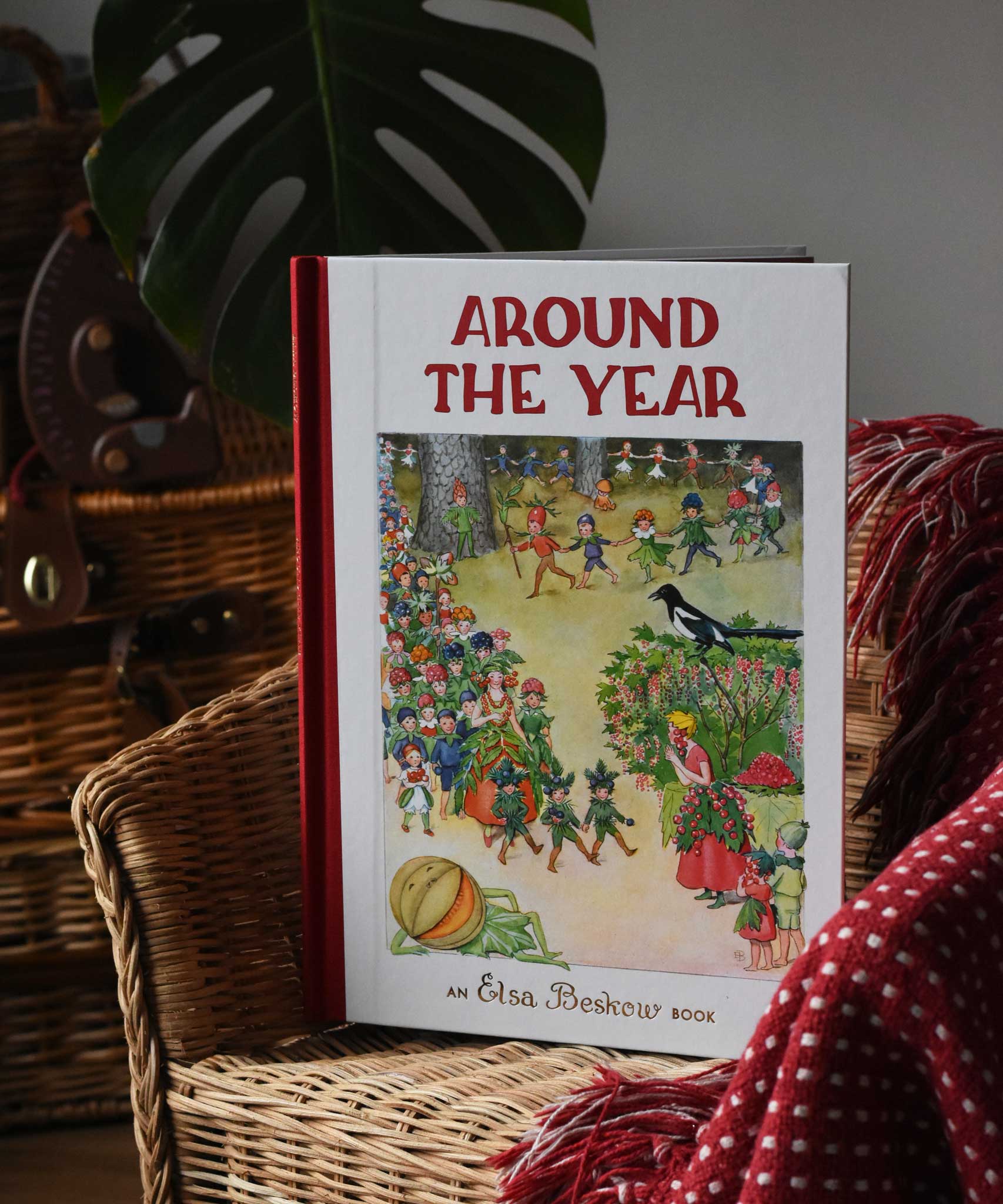 Around The Year children's hardback book by Elsa Beskow placed on a child's rattan chair next to a red blanket.