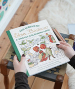A person holds a hardcover coloring book titled "The World of Elsa Beskow: A Vintage Colouring Book" over a woven stool in a home setting. The book cover features colorful vintage illustrations of strawberries, birds, and figures.