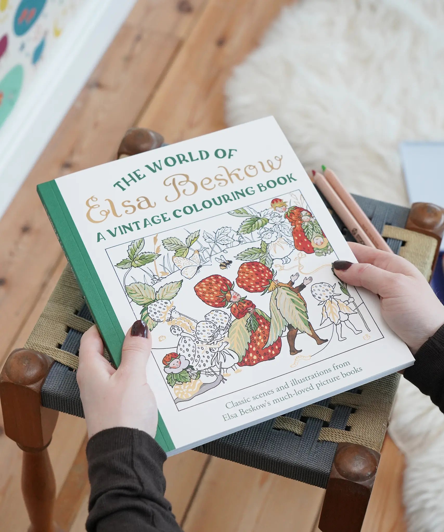 A person holds a hardcover coloring book titled "The World of Elsa Beskow: A Vintage Colouring Book" over a woven stool in a home setting. The book cover features colorful vintage illustrations of strawberries, birds, and figures.