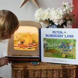 Peter in Blueberry Land children's hardback book by Elsa Beskow placed on a wicker hamper next to a Toverlux lamp with coordinating artwork. A child is looking at the toverlix silhouette art. 