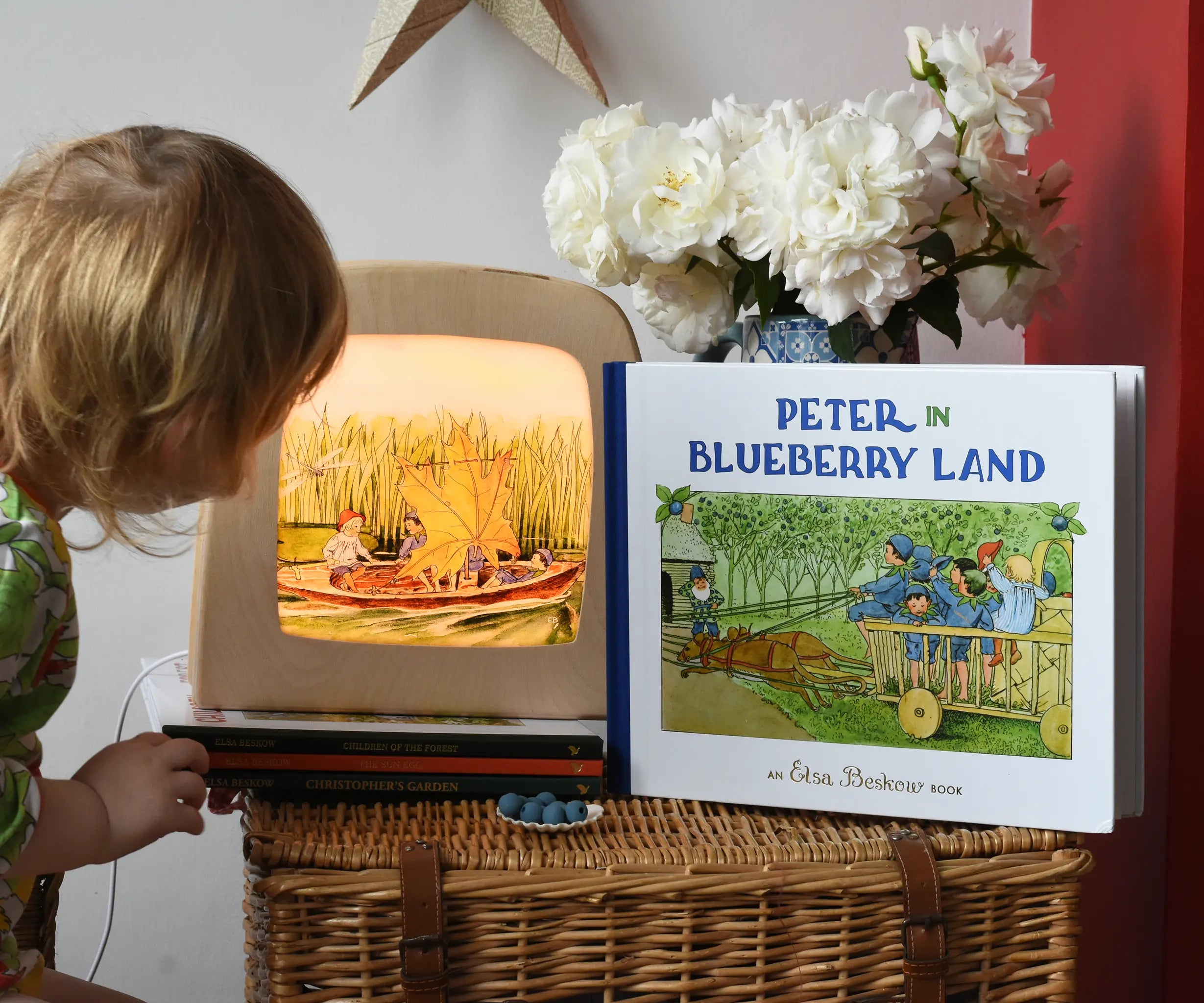 Peter in Blueberry Land children's hardback book by Elsa Beskow placed on a wicker hamper next to a Toverlux lamp with coordinating artwork. A child is looking at the toverlix silhouette art. 