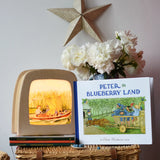 Peter in Blueberry Land children's hardback book by Elsa Beskow placed on a wicker hamper next to a Toverlux lamp with coordinating artwork.